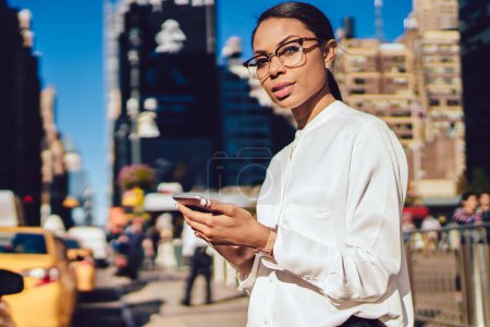 Téléchargez les photos : Portrait en demi-longueur d'une femme confiante et fière ceo en lunettes d'optique regardant la caméra en attendant le taxi en milieu urbain, une femme intelligente en tenue élégante tenant un téléphone cellulaire moderne - en image libre de droit