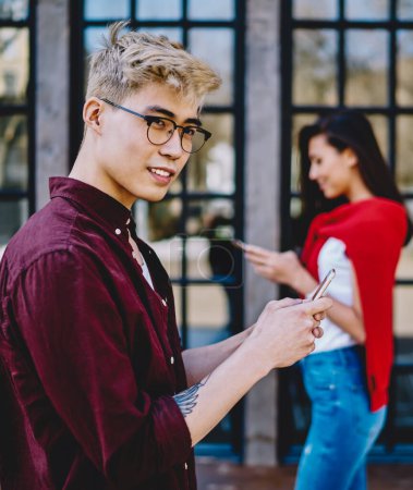 Photo for Handsome chinese hipster guy in eyeglasses looking away while messaging on smartphone and using 4G internet for communication with friend which standing on blurred background with cellular in hand - Royalty Free Image