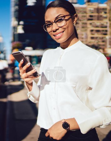 Téléchargez les photos : Portrait en demi-longueur d'une femme joyeuse dans des lunettes optiques pour protéger les yeux tenant un gadget pour smartphone et suivant les gps via une application en attendant la cabine jaune près de la route avec la circulation - en image libre de droit