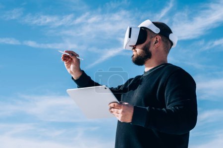 Foto de El arquitecto masculino milenario diseña la estructura de la nueva ciudad utilizando tabletas y auriculares de realidad virtual que crean un proyecto de dibujos electrónicos. Tecnología innovadora, masculina que toca el mundo VR con lápiz digital - Imagen libre de derechos