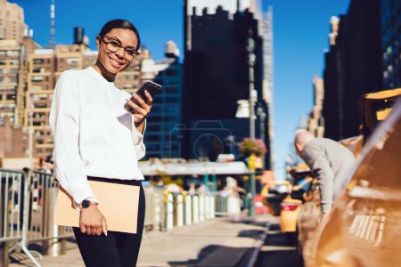 Téléchargez les photos : Portrait d'une jeune afro-américaine joyeuse aux lunettes froides souriant de conversations amusantes avec des amis via cellulaire, une fille positive regardant la caméra alors qu'elle se trouve dans la rue près de cabines jaunes - en image libre de droit