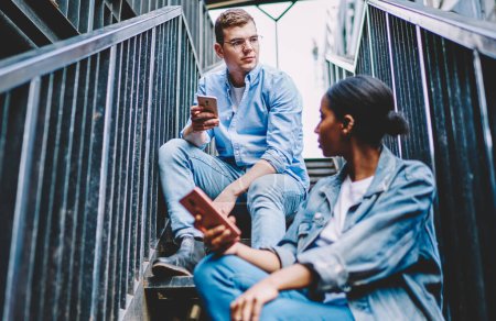 Photo for Millennial generation using modern smartphones for communication in social networks via public 4G internet connection.Multicultural young man and woman with mobile phones sitting on stairs of house - Royalty Free Image