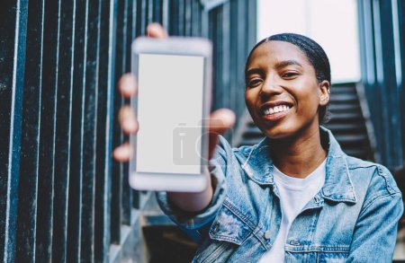 Photo for African American millennial female showing smartphone with mockup copy space screen for paste advertising on web content.Cheerful and happy dark skinned blogger holding in hand mobile phone presenting - Royalty Free Image