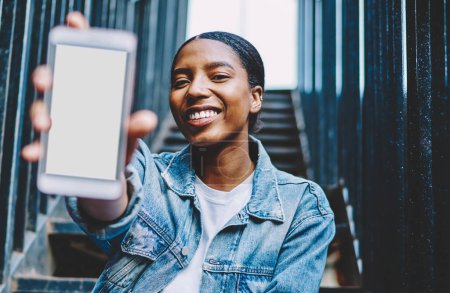 Photo for African American millennial female showing smartphone with mockup copy space screen for paste advertising on web content.Cheerful and happy dark skinned blogger holding in hand mobile phone presenting - Royalty Free Image