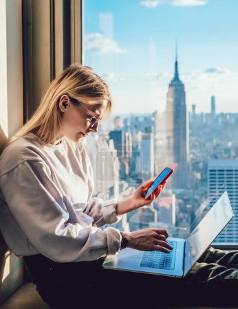Photo for Millennial hipster girl typing text on laptop keyboard while holding smartphone in another hand.Female traveler working on freelance with scenery view of New York downtown with famous Empire landmark - Royalty Free Image
