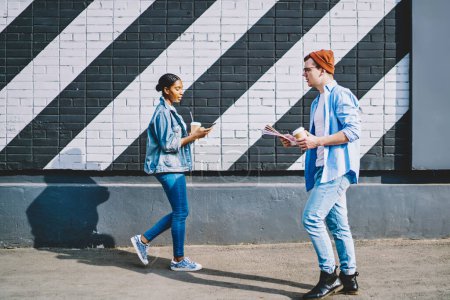 Photo for Multicultural hipster guys dressed in casual wear walking with coffee and chatting in social networks on smartphones.Addicted diverse young man and woman bloggers strolling with cellulars on street - Royalty Free Image