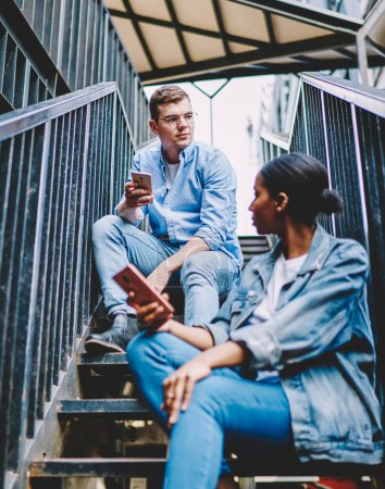 Photo for Millennial generation using modern smartphones for communication in social networks via public 4G internet connection.Multicultural young man and woman with mobile phones sitting on stairs of house - Royalty Free Image