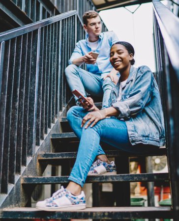 Photo for Portrait of cheerful african american young woman smiling at camera while updating profile on app on smartphone sitting with caucasian friend on stairs.Casual dressed diverse hipster guys with phones - Royalty Free Image