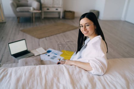 Photo for From above of smiling young woman in glasses and white shirt with dark hair working online on computer with financial documents - Royalty Free Image