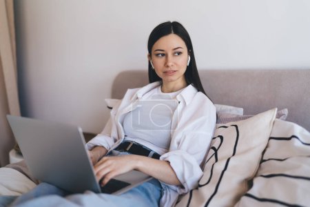 Photo for Young female freelancer in casual clothes and earbuds sitting on bed typing information on netbook during remote work from home while looking away thoughtfully - Royalty Free Image