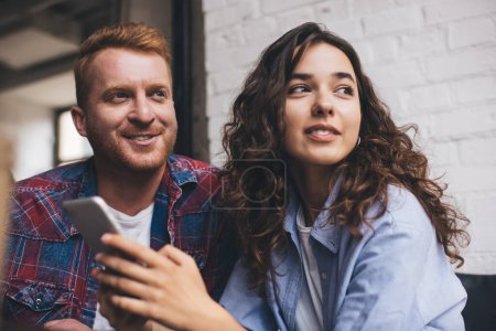 Photo for Caucasian male and female with cellular gadget looking away during leisure time, millennial best friends with smartphone technology thinking and smiling during date meeting in cafe interior - Royalty Free Image