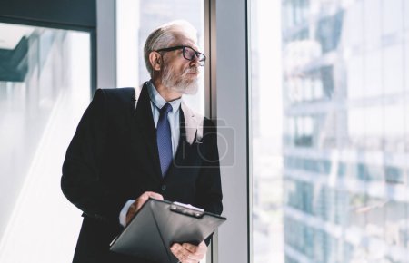 Pensive elderly businessman in elegant suit and eyeglasses looking away while leaning on iron bar supporting glass wall in modern office and holding clipboard in hand