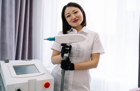 Foto de Especialista femenina positiva en guantes negros y uniforme blanco de pie en clínica moderna con láser de neodimio mientras se prepara para el procedimiento - Imagen libre de derechos