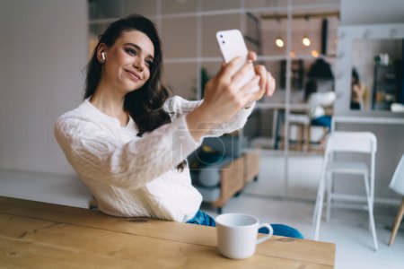 Photo for Positive young female in casual clothes smiling and taking self portrait on smartphone while sitting at table with cup of coffee - Royalty Free Image