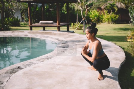 Photo for Full body of calm young ethnic barefoot female in activewear practicing Goddess pose with namaste hands and closed eyes on poolside - Royalty Free Image
