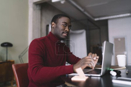 Photo for Serious black male freelancer in red sweater typing on laptop and looking at camera while working remotely from home office - Royalty Free Image