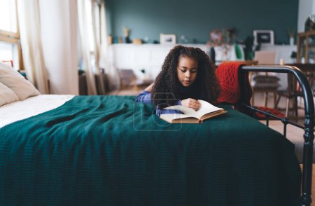 Téléchargez les photos : Concentré fille mignonne avec sombre bouclé livre de lecture de cheveux tout en étant couché sur le lit dans une chambre confortable lumineuse avec couverture verte et oreiller sur fond flou à la lumière du jour - en image libre de droit