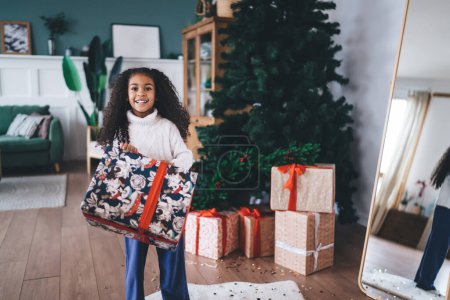 Photo for Pensive curly haired cute black little girl in casual clothes carrying wrapped Christmas gift box while standing in living room with Christmas tree and gift boxes while looking at camera - Royalty Free Image