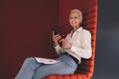 Téléchargez les photos : Femme d'affaires caucasienne senior en lunettes élégantes, utilisant un smartphone et tenant des documents, confortablement inclinée dans une chaise à gousse rouge chic, reflétant la flexibilité du travail et l'engagement numérique senior - en image libre de droit