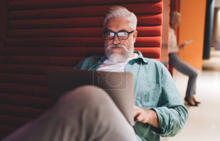 Téléchargez les photos : Homme âgé concentré avec barbe grise et lunettes travaillant sur un ordinateur portable dans un salon moderne avec panneaux acoustiques rouges, illustrant la capacité d'adaptation des entrepreneurs plus âgés aux espaces de travail innovants - en image libre de droit