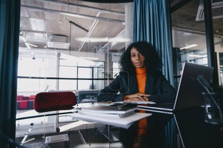 Photo for Serious Black businesswoman, wearing stylish blazer and vibrant orange sweater, sits at office desk filled with documents, contemplating while looking at the camera, in a well-lit modern workspace. - Royalty Free Image