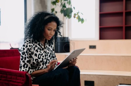 Photo for Concentrated Black female financial trader in leopard print blouse analyzing real-time data on tablet in a stylish office, with a touch of executive flair - Royalty Free Image