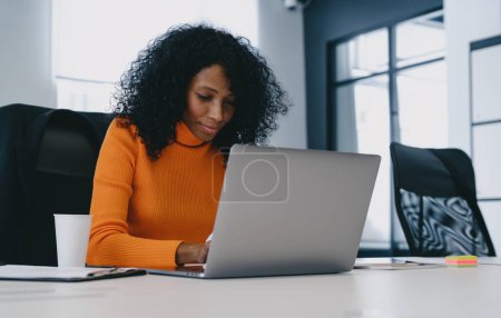 Konzentrierte professionelle Frau in leuchtend orangefarbenem Top greift in einem modernen Büro zum Laptop und verkörpert fokussierte Produktivität.