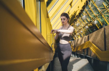 Foto de Enfocada joven mujer caucásica en una camiseta deportiva de color topo y polainas negras ajustando su reloj de fitness en un día soleado en las escaleras amarillas urbanas - Imagen libre de derechos
