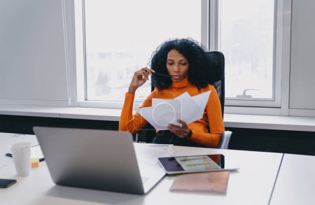 Analytische afroamerikanische Finanzanalystin in ihren Dreißigern, die einen Finanzbericht in einem modernen Co-Working-Raum kritisch bewertet, mit Laptop und Tablet auf dem Schreibtisch, ihr orangefarbener Rollkragen deutet auf