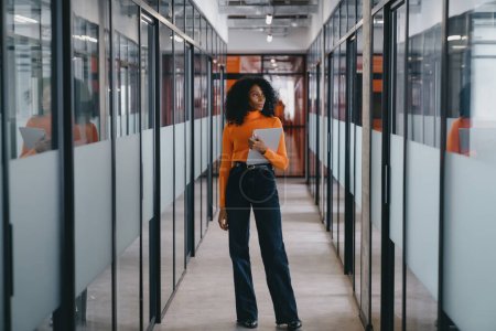 Téléchargez les photos : Femme d'affaires noire en haut orange et pantalon noir marchant avec caméra dans le couloir de bureau moderne, affichant un style de vie professionnel créatif. - en image libre de droit