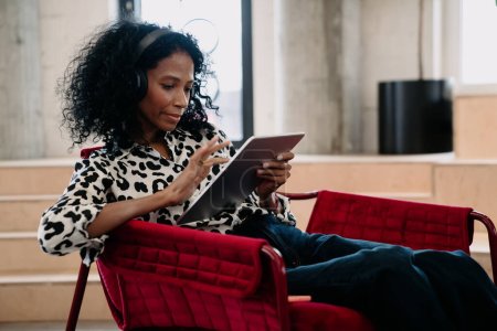 Téléchargez les photos : Engrossed Black expert en finance féminine avec écouteurs utilise tablette pour les études de marché dans le bureau moderne, mettant en valeur les prouesses analytiques et la compétence numérique - en image libre de droit