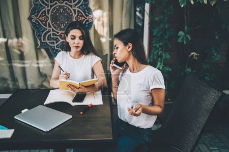 Photo for Concentrated young businesswoman in white t shirt speaking on smartphone and colleague holding notebook and pen while sitting together during informal meeting in cafeteria - Royalty Free Image