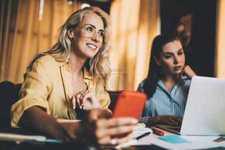 Cheerful young blond female colleague in glasses using smartphone while sitting next to focused coworker with laptop during informal meeting in flat