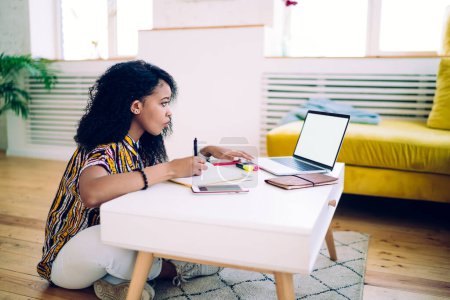 Vista lateral de la mujer étnica moderna adulta en traje casual sentado en la mesa en la escritura del piso en el cuaderno mientras se trabaja en el ordenador portátil con pantalla en blanco en casa 