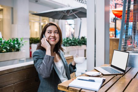 Happy Asian businesswoman dressed in formal suit sitting in front of laptop with cup of coffee in hand and looking away during phone conversation