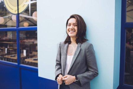 Bastante alegre joven mujer asiática en chaqueta casual de pie y apoyado contra la pared azul de la cafetería mientras sonríe y mira hacia otro lado