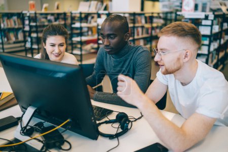 Junge fröhliche intelligente Studenten bereiten gemeinsam ein Projekt vor und suchen Informationen am Computer in der großen Universitätsbibliothek, während sie Projektideen diskutieren
