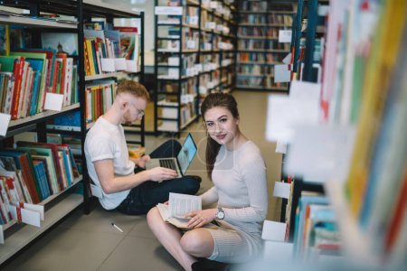 Young smiling attractive woman sitting and flipping pages of book looking at camera while serious male student nearby browsing laptop among library shelves