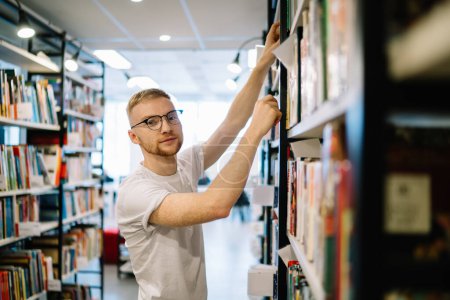 Téléchargez les photos : Vue latérale du calme heureux homme barbu adulte en chemise blanche et lunettes regardant la caméra tout en choisissant livre sur l'étagère dans la bibliothèque de lumière - en image libre de droit