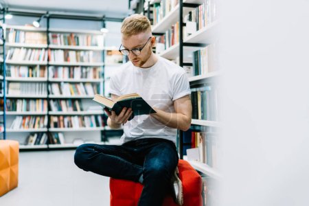 Téléchargez les photos : Homme barbu adulte pensif en vêtements décontractés et lunettes assis jambes croisées sur coulf rouge et livre de lecture près des étagères dans la bibliothèque moderne lumière - en image libre de droit