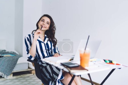 Smiling elegant adult lady holding pen and touching lips while sitting at desk with laptop phone stationery and to go glass with orange juice against white wall in light bedroom