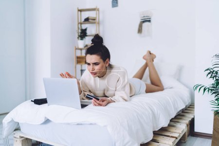 Confused pretty adult female in casual clothes holding credit card and focusing on screen while trying using laptop for online shopping during lying in bed in light apartment on blurred background