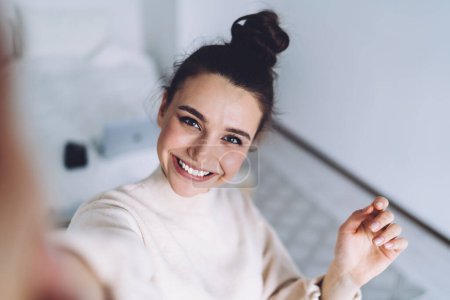 Hübsche junge Frau mit breitem Lächeln steht nach der Arbeit mit Laptop im Schlafzimmer und hat Spaß dabei, ein leichtes fröhliches Selfie auf verschwommenem Hintergrund zu machen