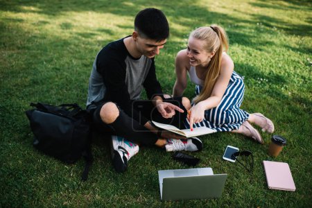 Téléchargez les photos : D'en haut souriant amis intelligents pointant vers livre ouvert tout en apprenant ensemble sur l'herbe dans le parc d'été après les cours universitaires - en image libre de droit