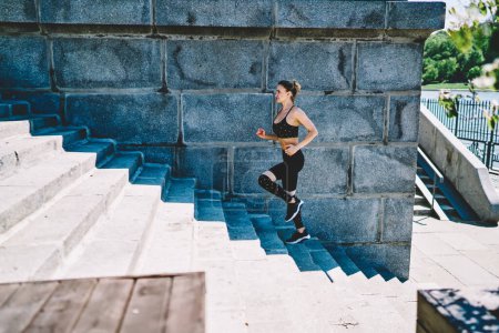 Téléchargez les photos : Vue latérale de la dame sportive mince en noir élégant vêtements de sport surmontant les escaliers à bascule rapide au stade en plein air le jour ensoleillé d'été - en image libre de droit