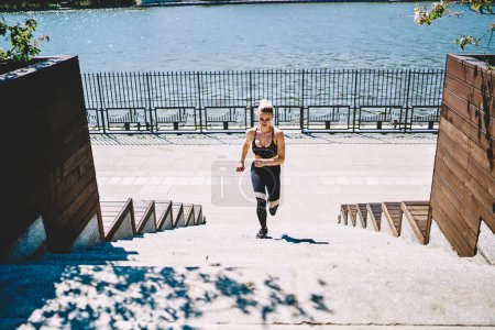 Téléchargez les photos : D'en haut de coureuse concentrée en vêtements de sport noirs couvrant la distance sur les escaliers à bascule du quai par une journée ensoleillée d'été - en image libre de droit