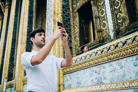 Photo for Serious spanish male travel blogger standing near golden temple making picture of asian notable places and architecture, man tourist shooting video for blog using smartphone camera outdoors - Royalty Free Image