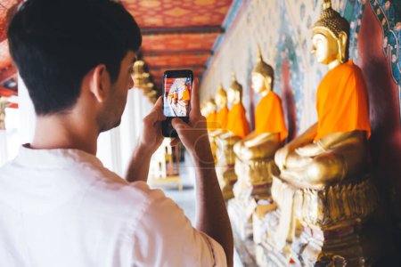 Photo for Back view of male blogger taking picture of buddha in asian temple during journey on free time, young millennial hipster guy using cellular camera for making content for social network in destination - Royalty Free Image