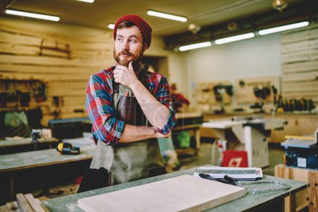 Ensamblador masculino creativo en elegante atuendo casual y delantal, pensando mientras comienza a trabajar con madera y mirando hacia otro lado en el taller 