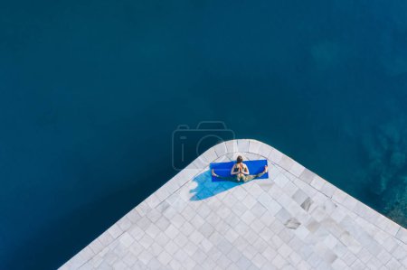 Foto de Vista superior de la mujer delgada en ropa deportiva sentada sobre cordel en la estera de yoga cerca del agua y estirándose durante el entrenamiento matutino - Imagen libre de derechos